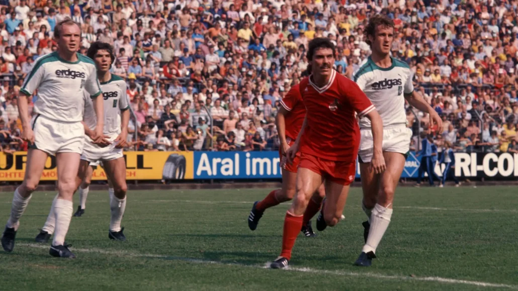 1976 die Version mit V-Ausschnitt: Herbert Zimmermann (1. FC Köln) im Laufduell mit Rainer Bonhof von Borussia Mönchengladbach.